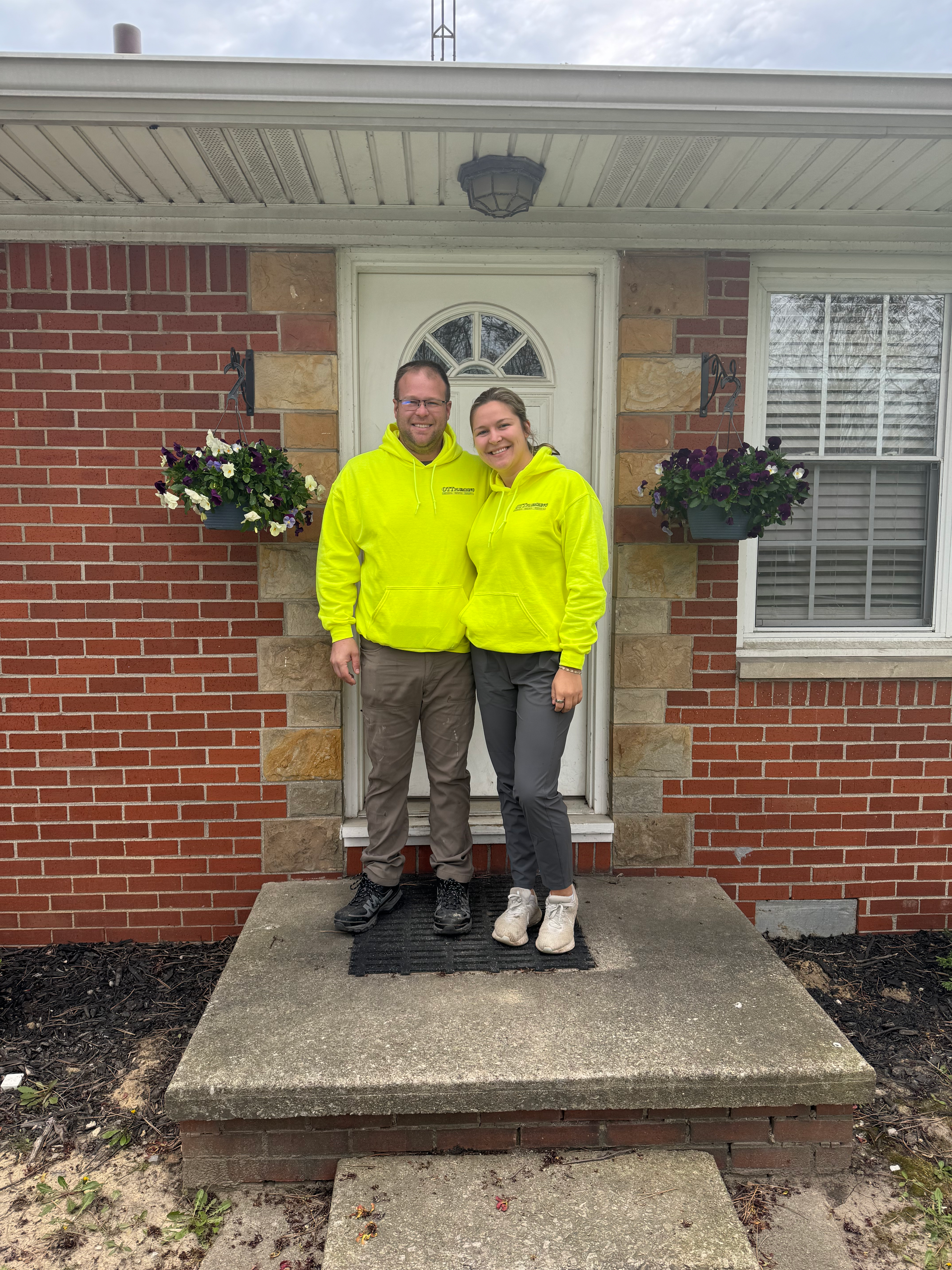 Curt and Cassie Ott in branded yellow Ott Plumbing jackets, smiling on the front porch of a Toledo brick home.